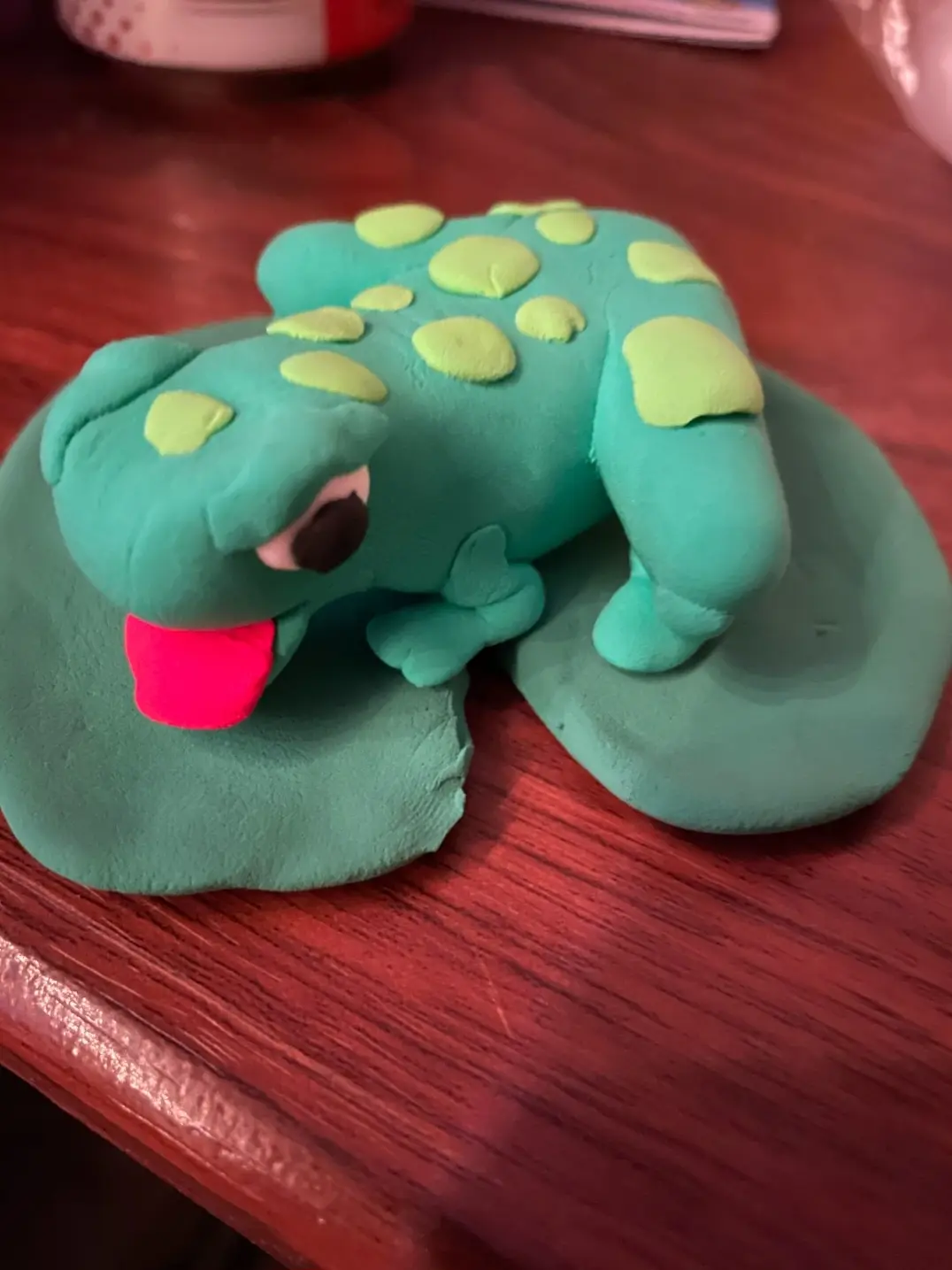A handmade clay frog with bright green spots and a pink tongue sticking out, sitting on a green clay lily pad on a wooden…