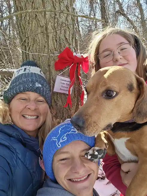 Three people and a dog pose for a selfie outdoors in winter near a tree with a sign reading 'Lamberton Lake Fen Nature…