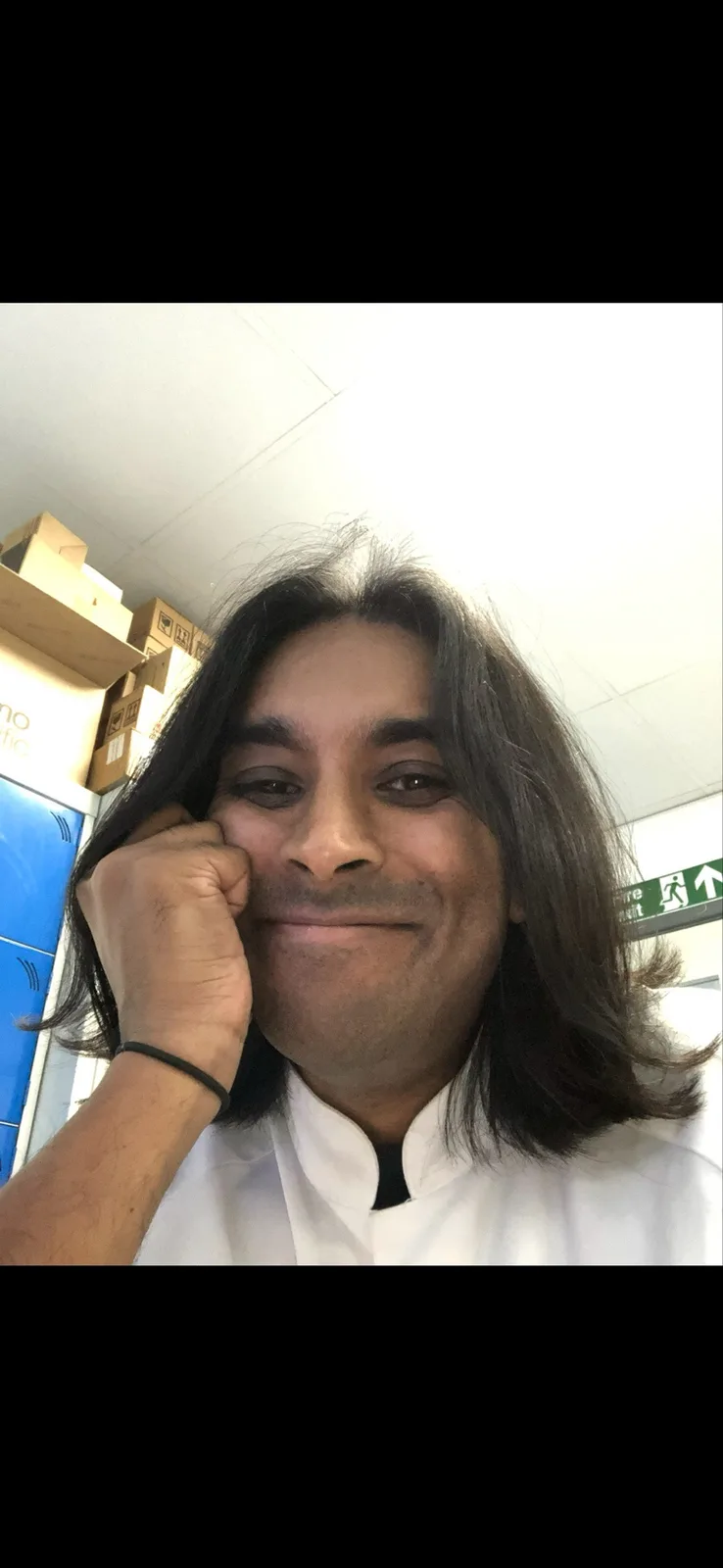 A man with long dark hair and a white collared shirt smiles while holding his hand to his cheek, standing in an indoor space…