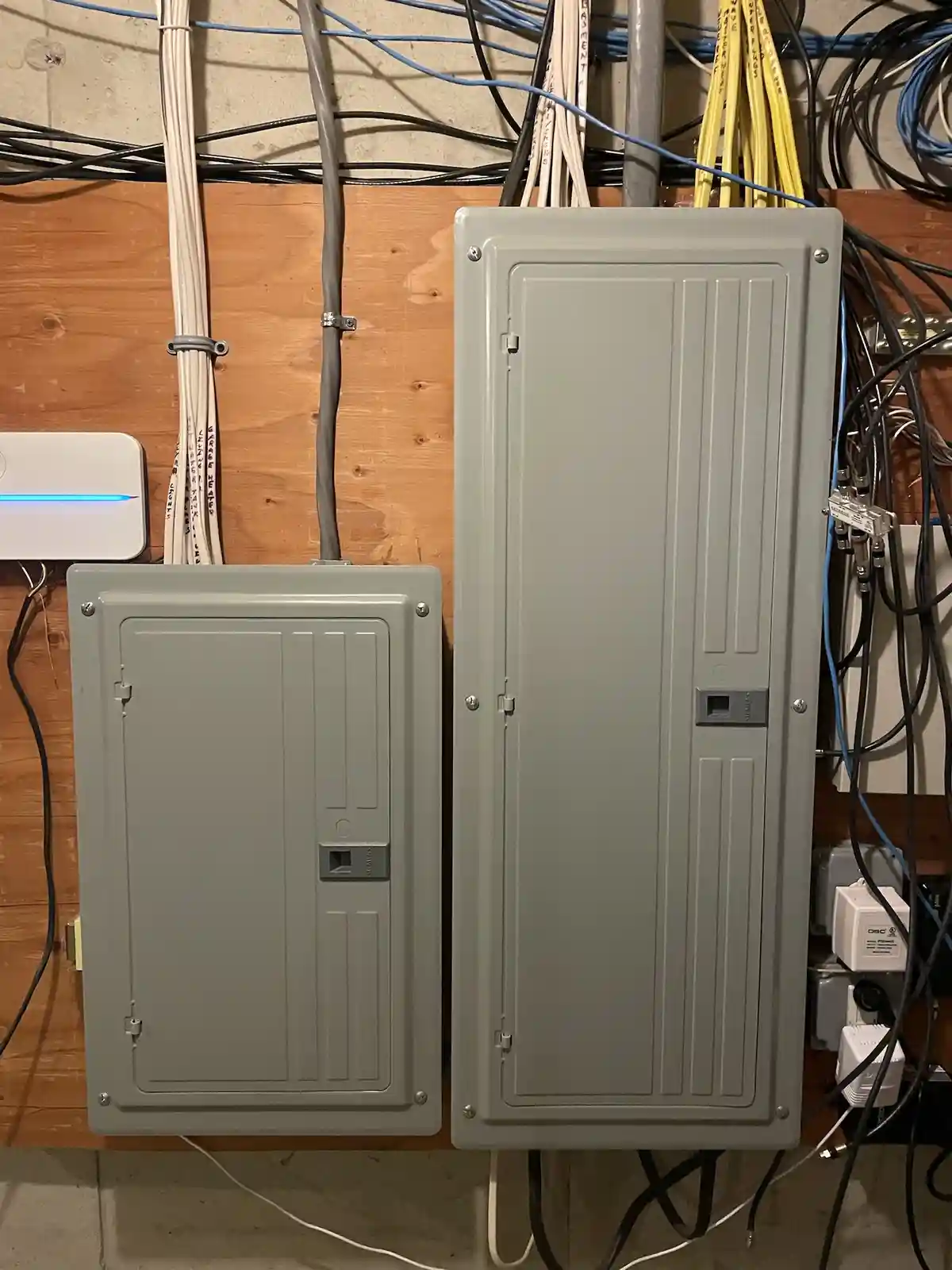 Two gray electrical panels mounted on a wooden wall surrounded by tangled wires and cables. The smaller panel is on the…