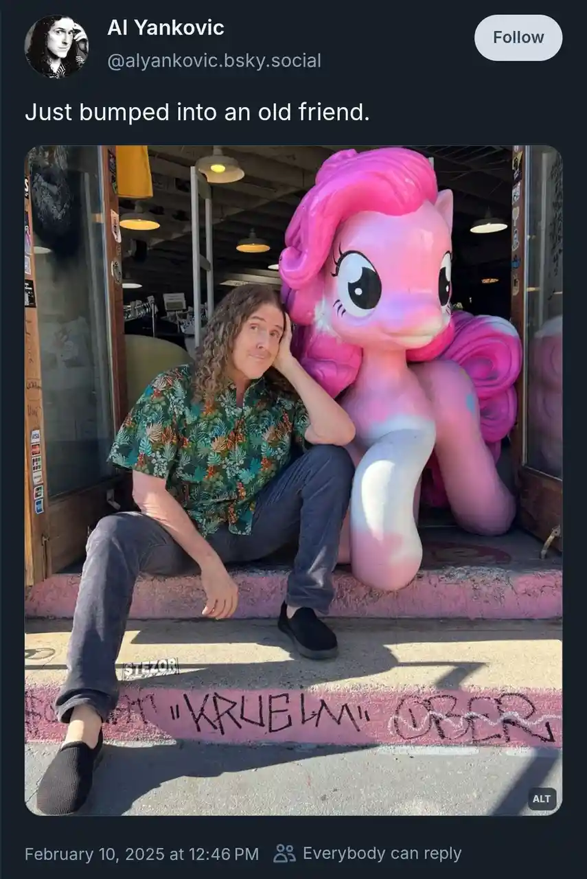 A man with long curly hair, wearing a patterned shirt and dark pants, sits on a pink-painted step next to a large, pink…