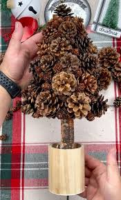 Two hands hold a small, handmade Christmas tree made entirely of pinecones arranged on a wooden base, set against a red and…