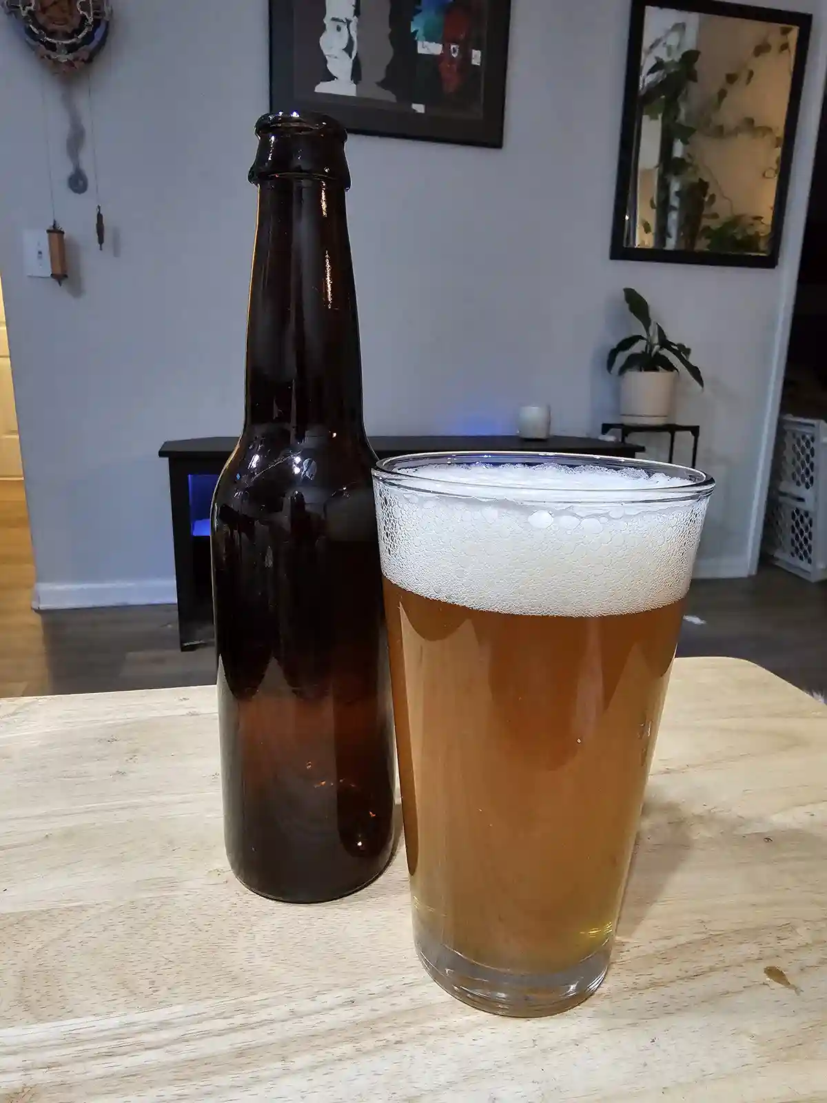 A dark brown beer bottle stands next to a tall glass filled with amber beer and a thick white head, both resting on a light…