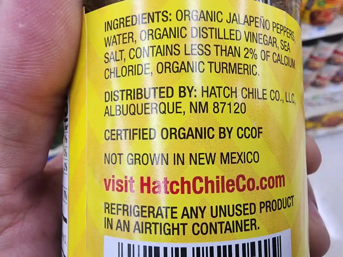 A close-up of a yellow-labeled jar of jalapeño pepper product held in a hand, showing its ingredient list and distributor…