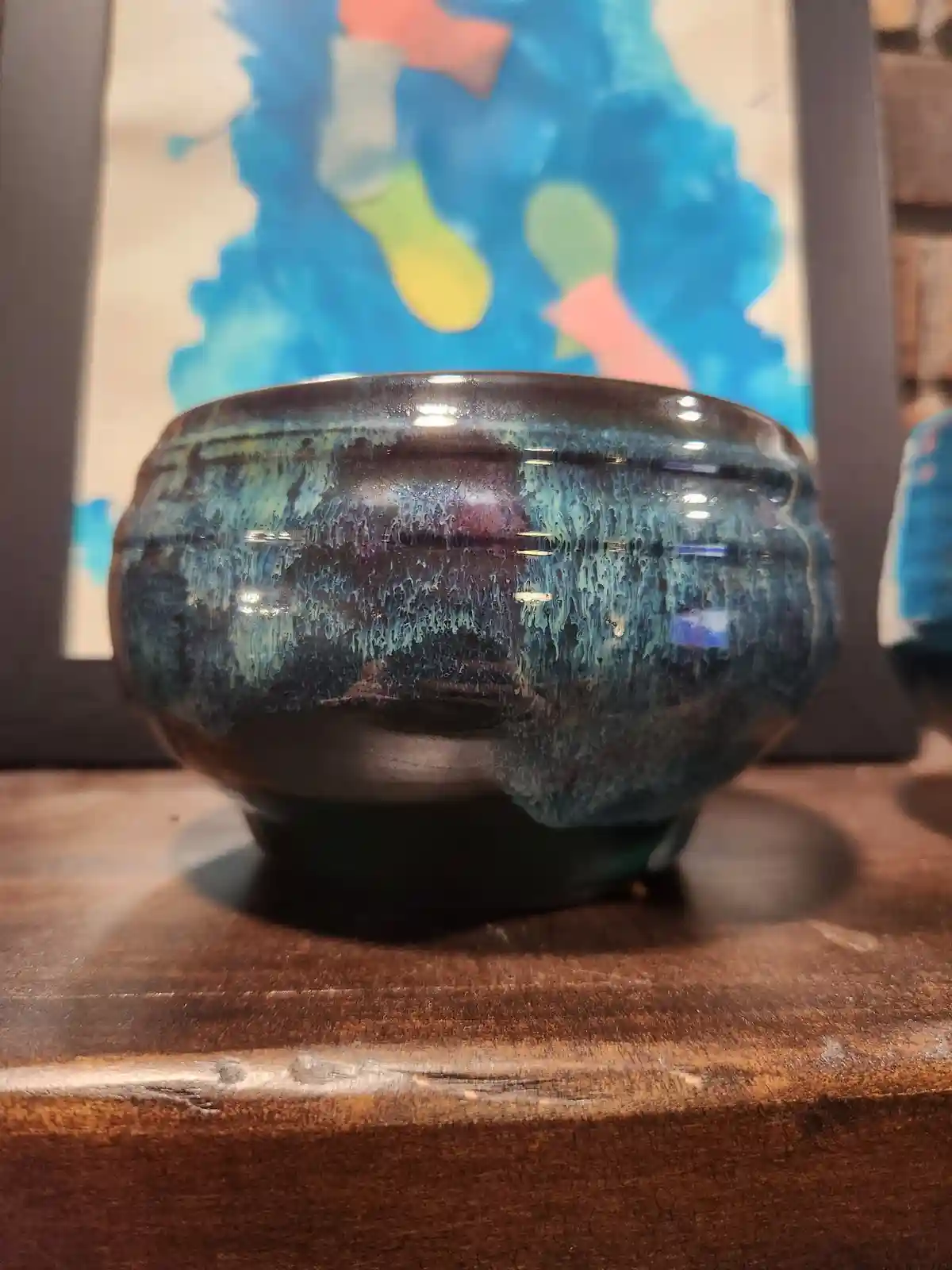 A glossy, dark blue ceramic bowl with streaks of teal and purple glaze sits on a wooden surface, with a colorful abstract…