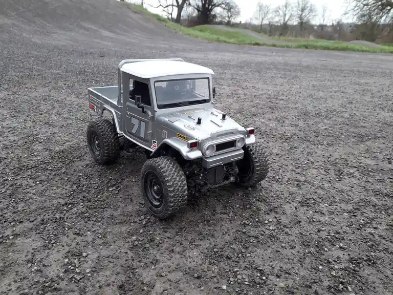 A silver remote-controlled model of a classic Japanese pickup truck, resembling a Toyota Land Cruiser FJ40, sits on a gravel…