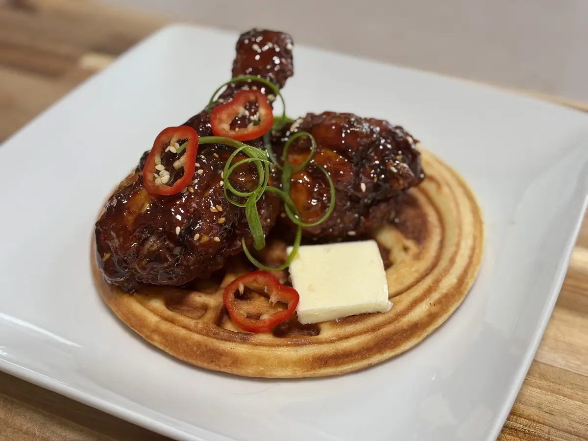 A close-up photo shows two glazed chicken legs resting on a golden-brown waffle on a white plate, garnished with sliced red…