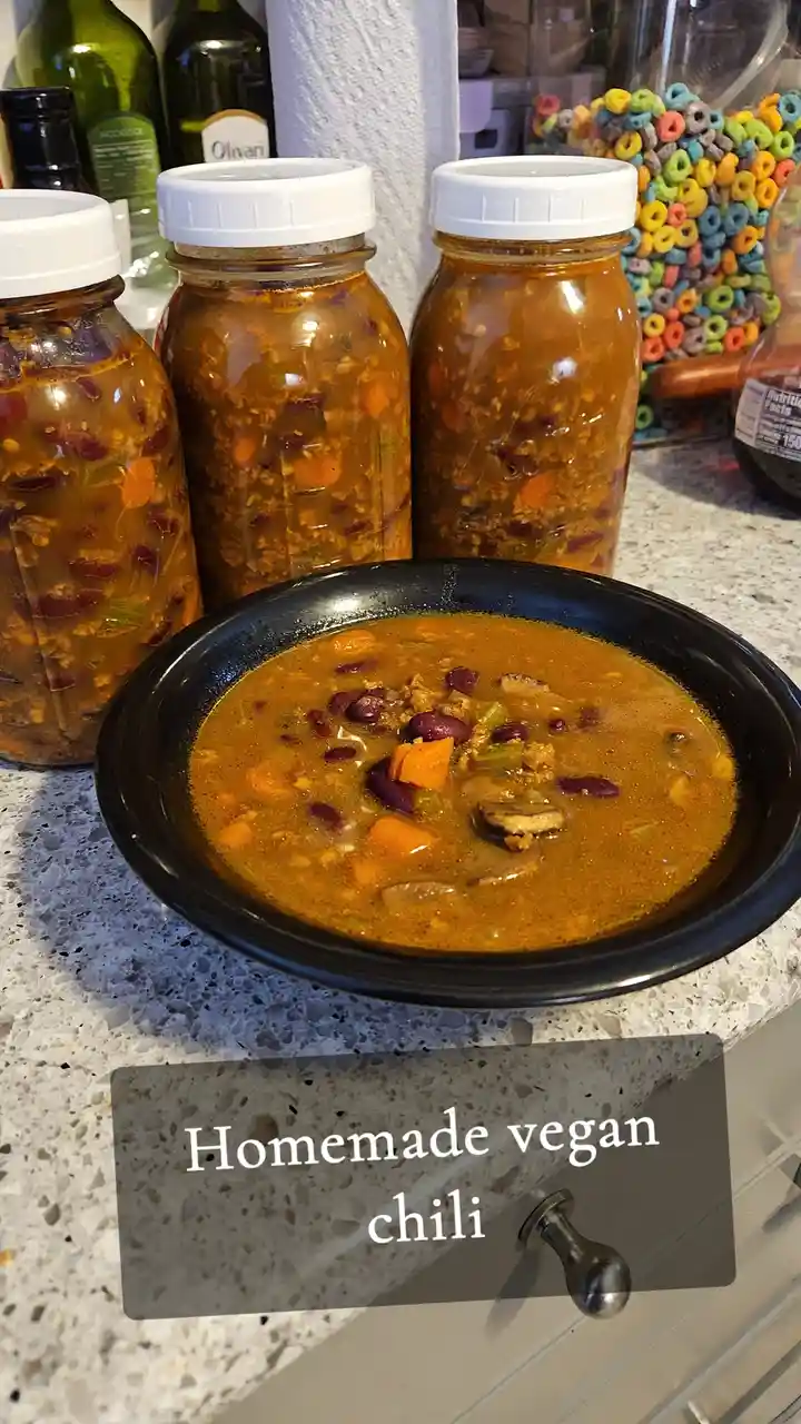 A black bowl of homemade vegan chili sits on a speckled countertop, with three glass jars filled with the same chili behind…
