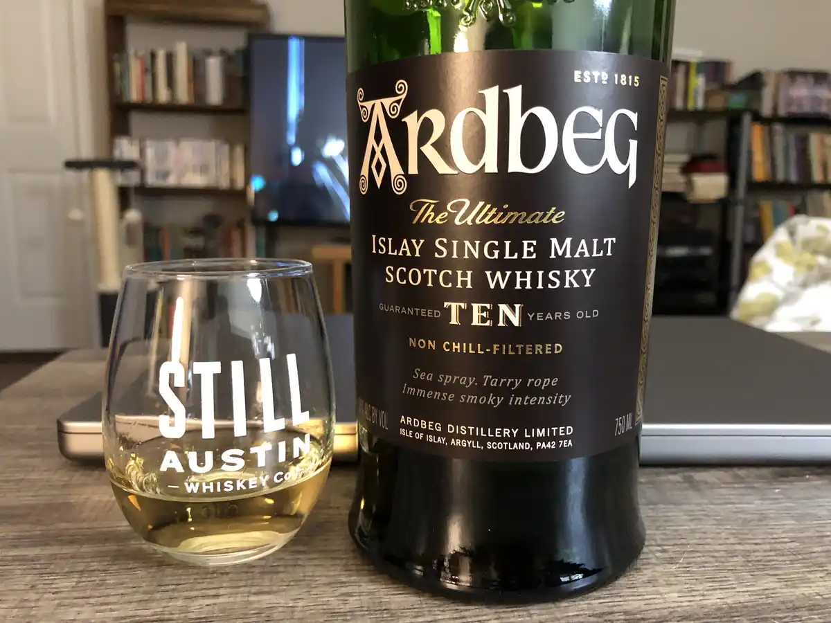A bottle of Ardbeg Ten Year Old Islay Single Malt Scotch Whisky sits next to a glass labeled 'STILL AUSTIN - WHISKEY CO'…