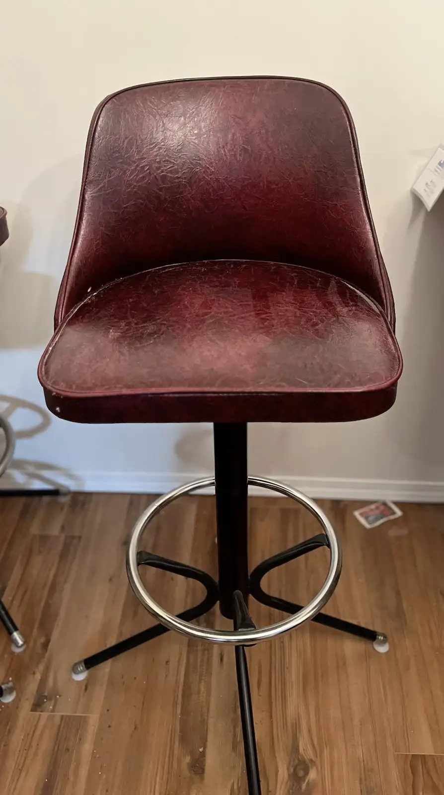 a close-up photo of a maroon, cracked faux-leather bar stool with a black metal base and chrome ring, standing on a wooden…