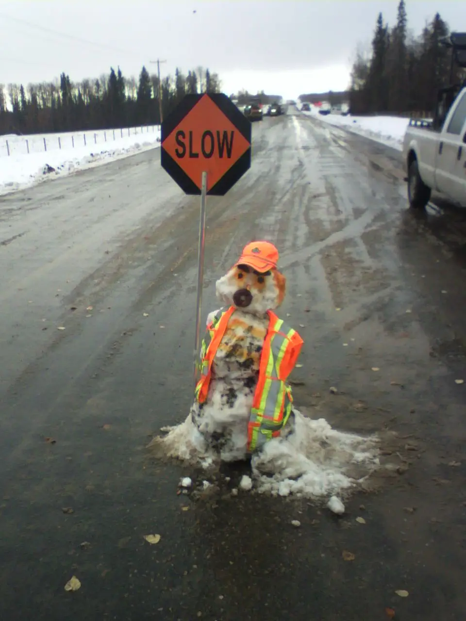 A snowman dressed in an orange construction vest and cap stands in the middle of a wet, snowy road, holding a 'SLOW' traffic…