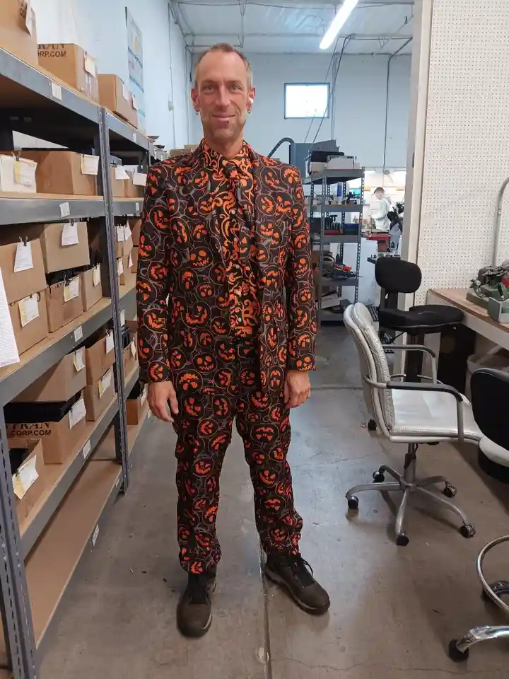 A man stands in a warehouse or workshop wearing a full-body suit covered in orange jack-o'-lantern faces on a black…