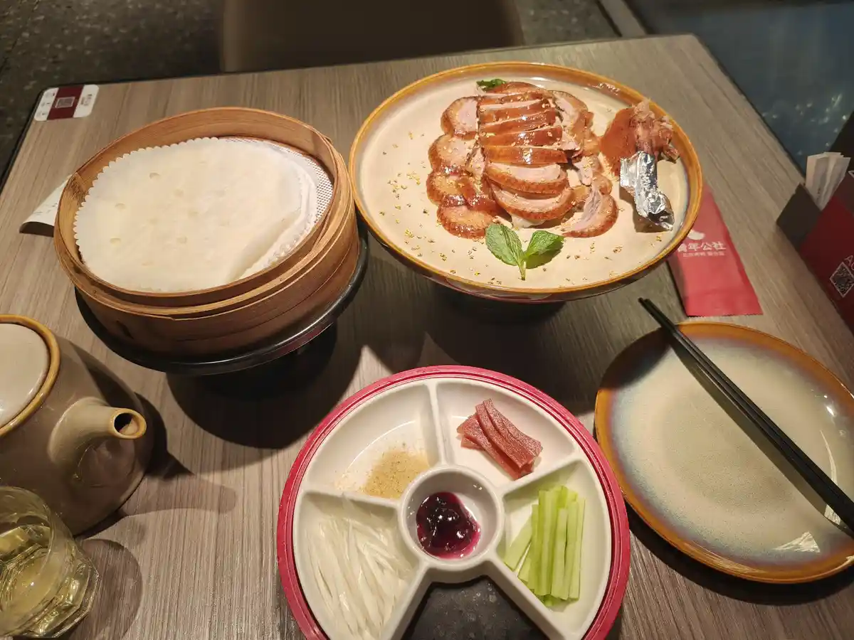 A table set with a full Beijing roast duck meal including sliced duck, steamed pancakes in a bamboo steamer, and a…