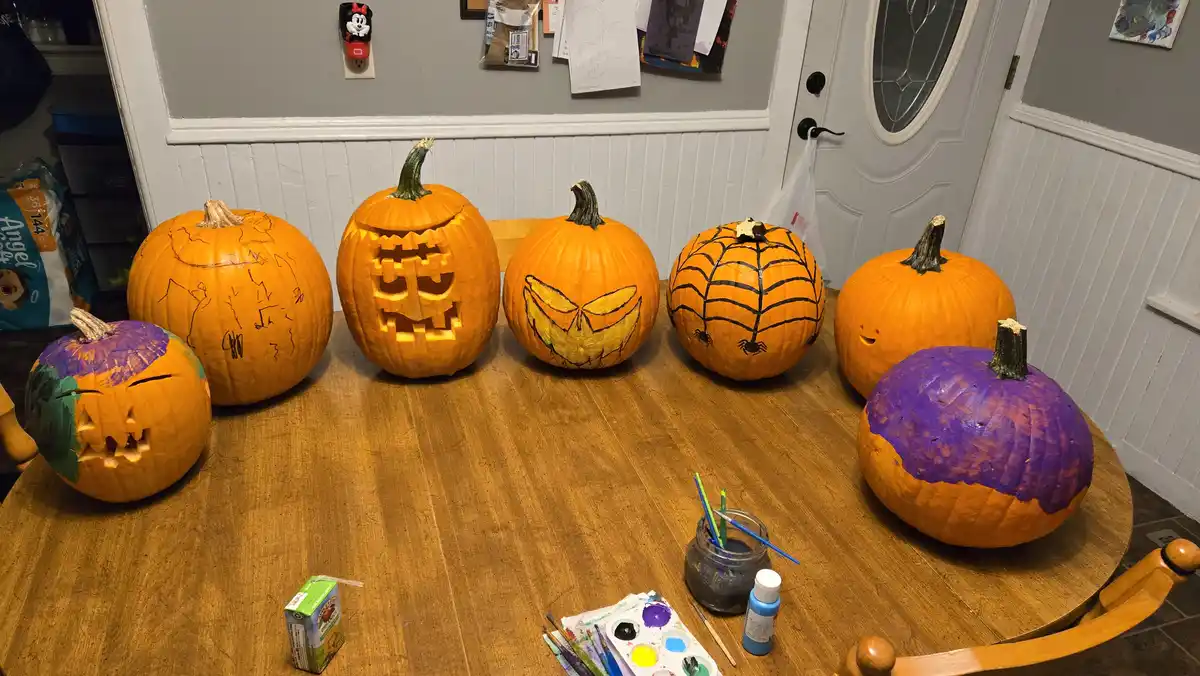 Seven pumpkins sit on a wooden table, varying in decoration from carved faces to painted spiderwebs and purple paint. Some…