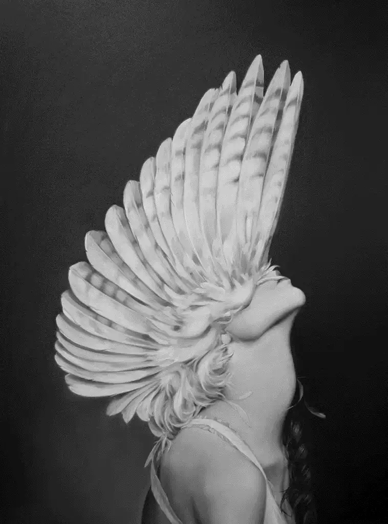 A black-and-white photo shows a woman in profile, wearing a large, elaborate feather headdress that fans out dramatically…