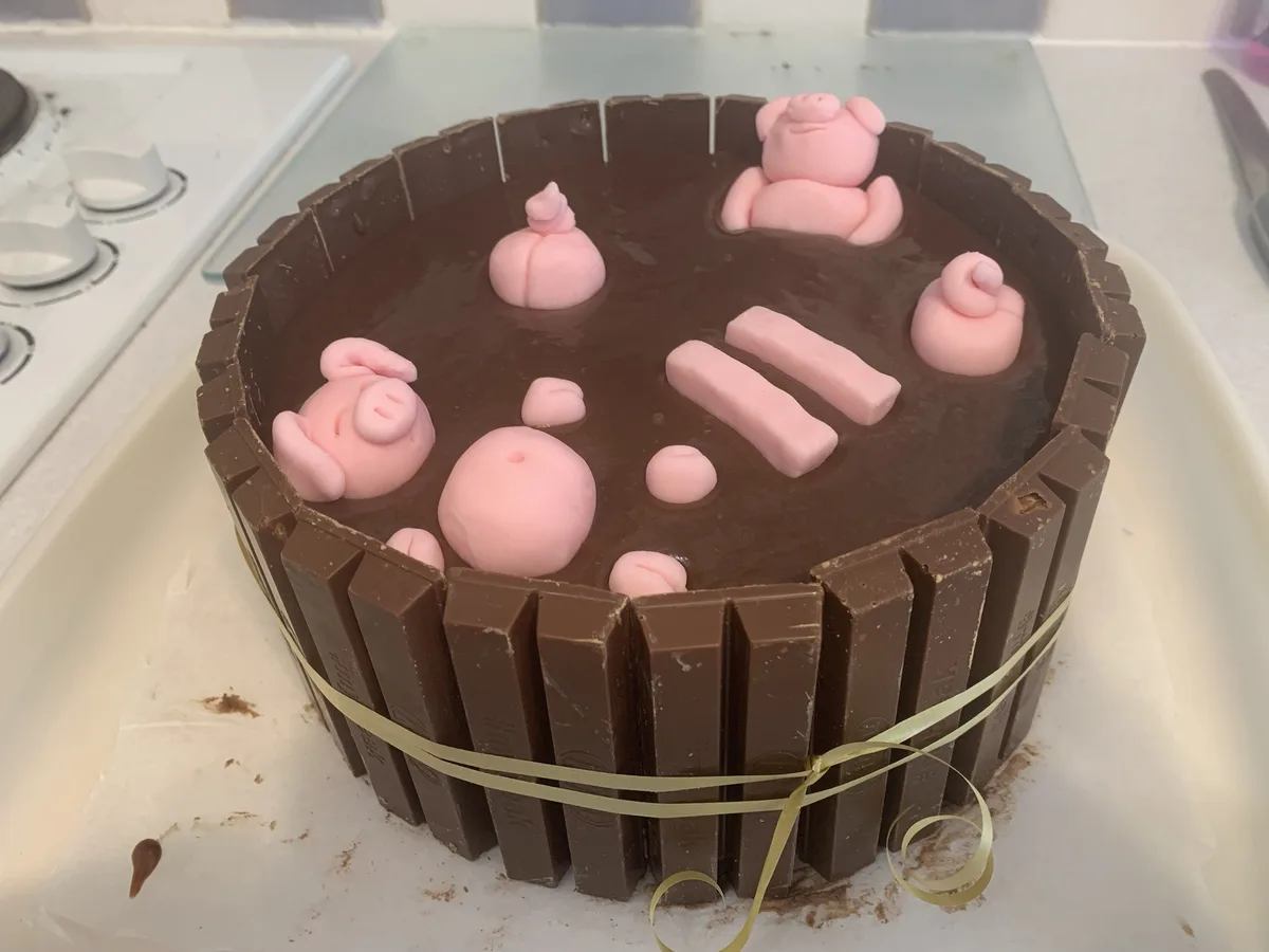 A round chocolate cake decorated with pink fondant figures resembling pigs and pig-related body parts, including a full pig…