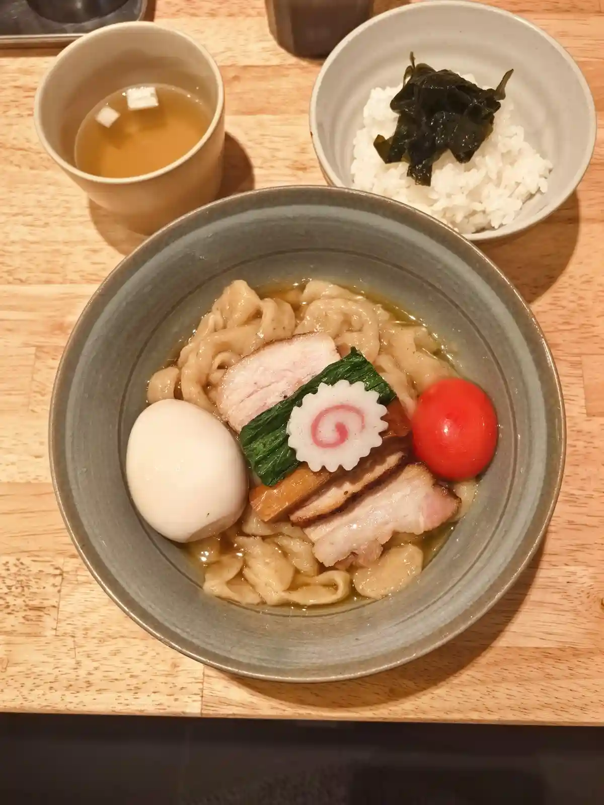 A top-down photo of a Japanese ramen bowl with udon noodles, sliced pork, a soft-boiled egg, a cherry tomato, and a…