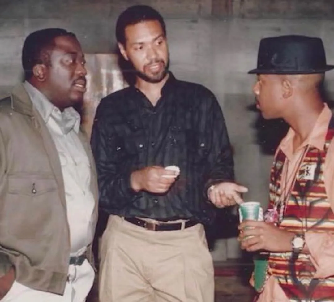 Three men are standing together in a dimly lit room. The man on the left has his mouth open as if speaking or reacting. The…