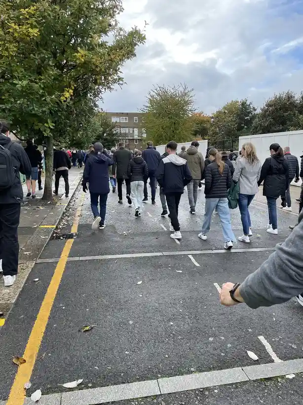 A group of people walks away from the camera down a paved road marked with parking lines and scattered autumn leaves. In the…