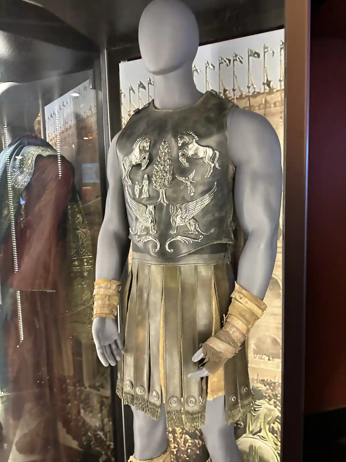 a gray mannequin displays an ornate, sleeveless Roman-style armor with silver embossed designs including two horses, a tree…