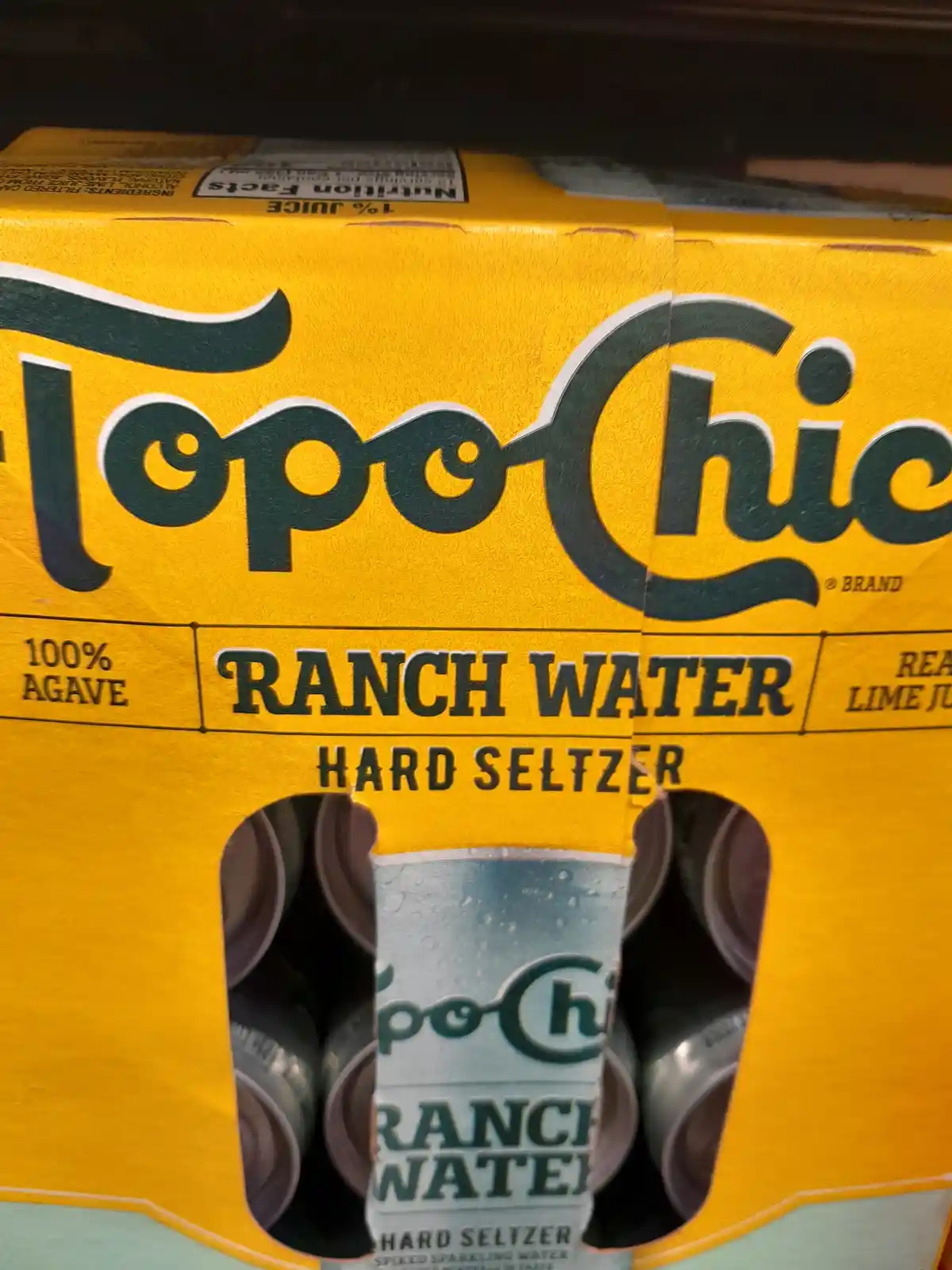 A close-up of a yellow Topo Chico Ranch Water hard seltzer six-pack box, with one can visible through the opening. The box…
