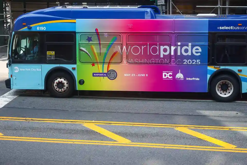 A New York City Bus, number 6190, is parked on a street with yellow road markings. Its side is covered in a vibrant rainbow…