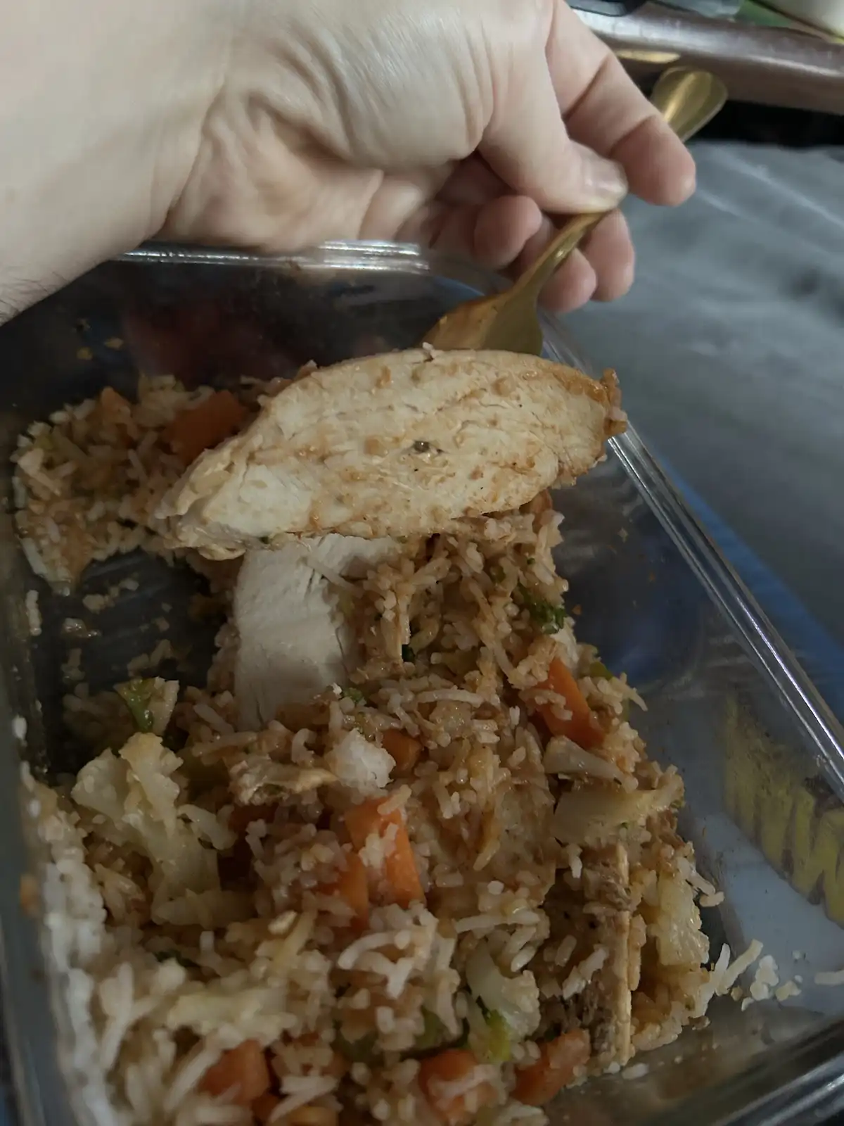 A hand holds a golden fork lifting a large slice of cooked chicken breast from a glass container filled with rice and mixed…
