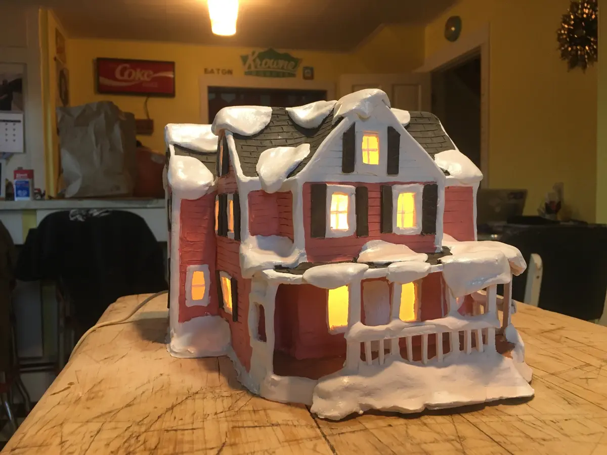 A handcrafted, illuminated gingerbread house model with white icing resembling snow on its roof and porch, sits on a wooden…