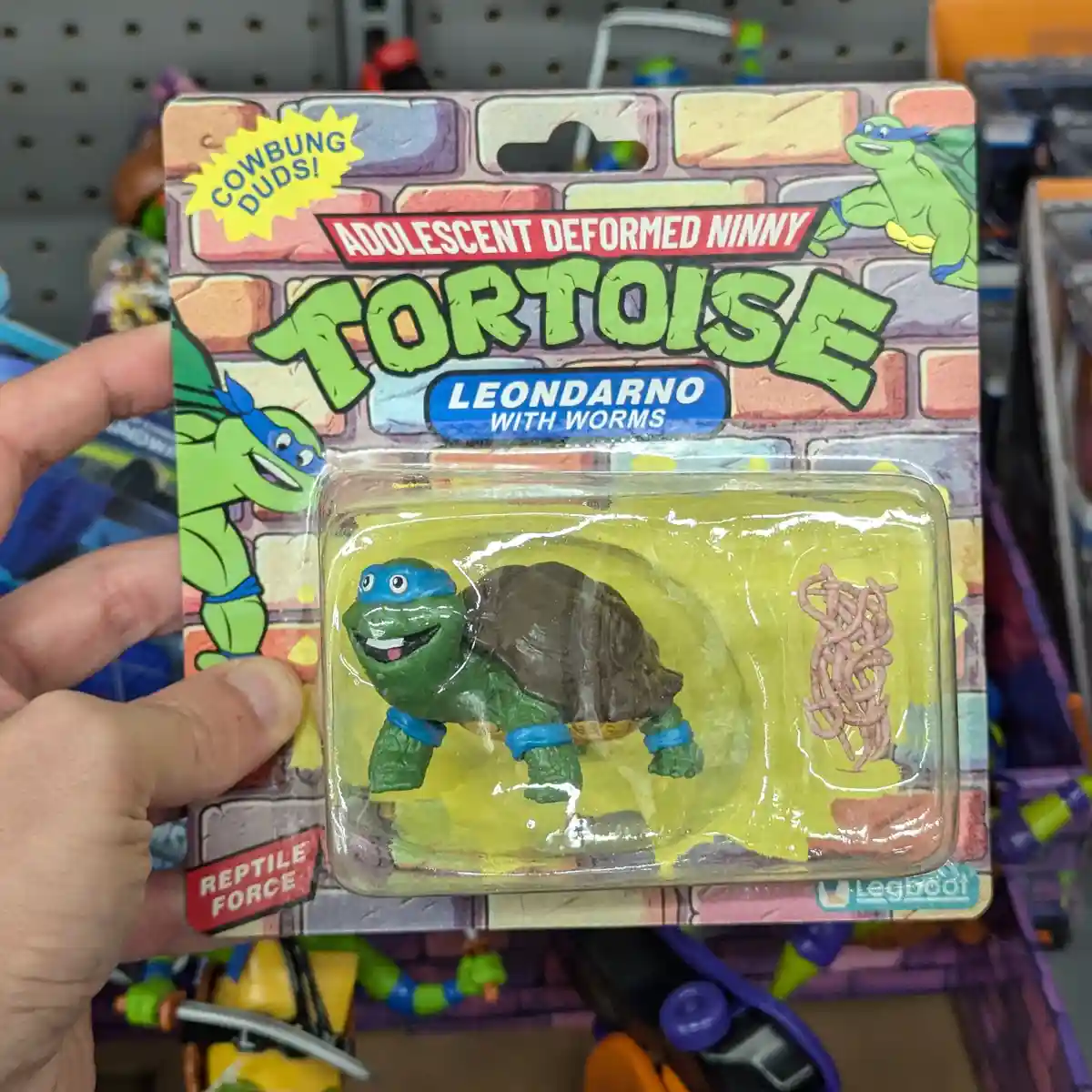 A hand holds a toy package parodying Teenage Mutant Ninja Turtles, featuring a green turtle figure with a blue bandana and a…