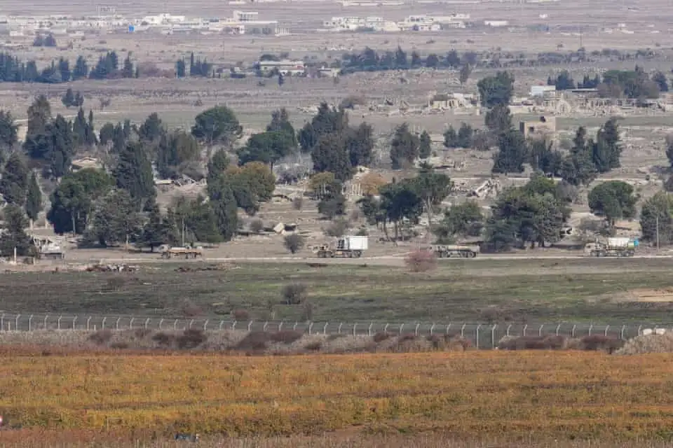 A wide landscape photo shows several Israeli military trucks driving along a dirt road through a rural area with trees…