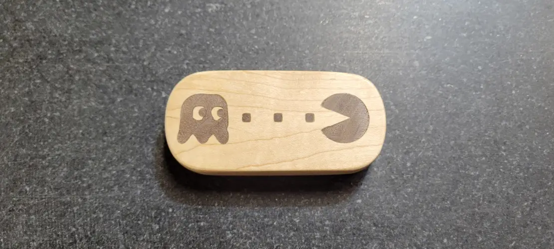 A light-colored wooden object, shaped like a pill, rests on a dark gray speckled surface. It features a laser-etched Pac-Man…