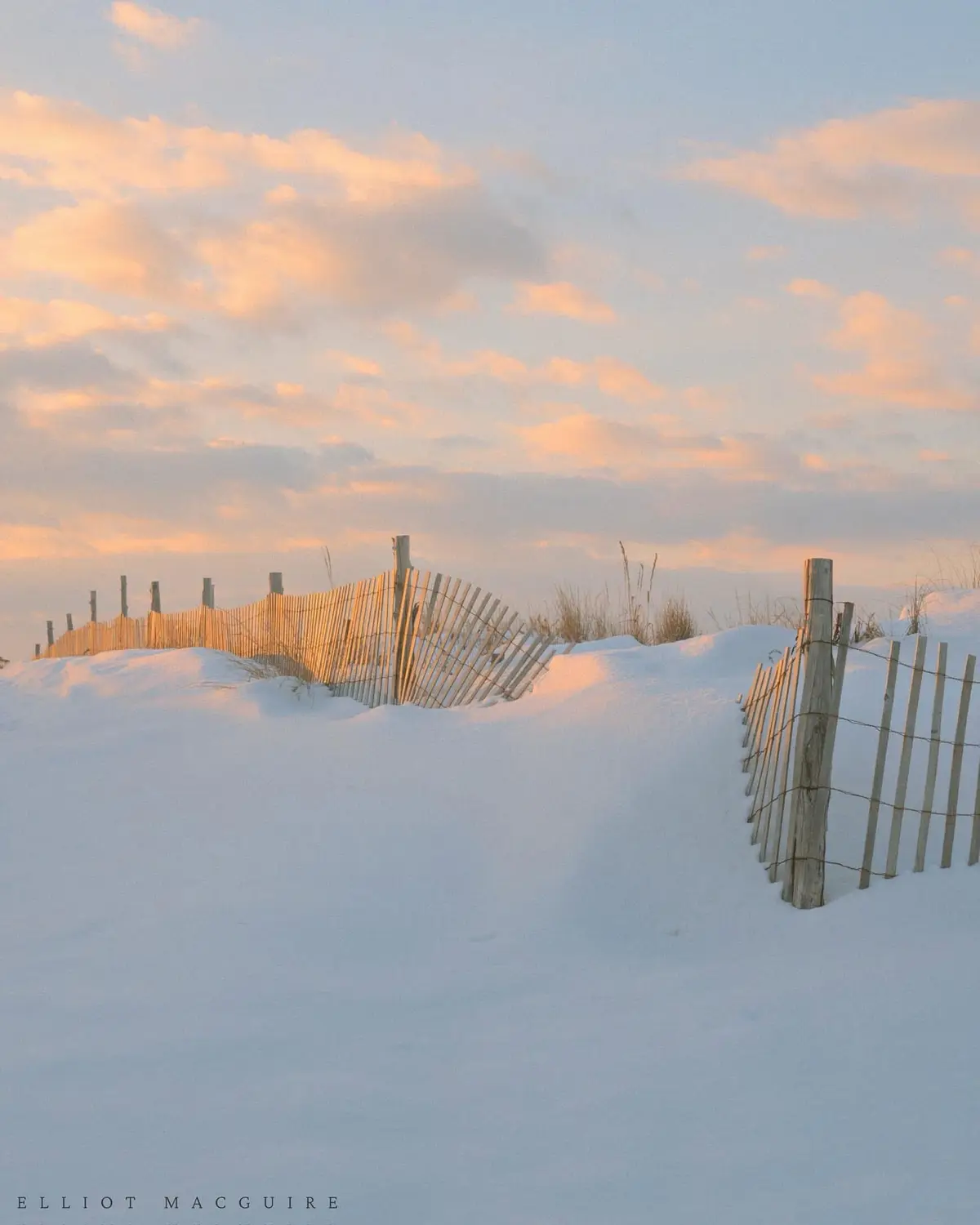 A serene winter scene shows a wooden beach fence partially buried in deep snow under a soft, pastel-colored sky at sunrise…