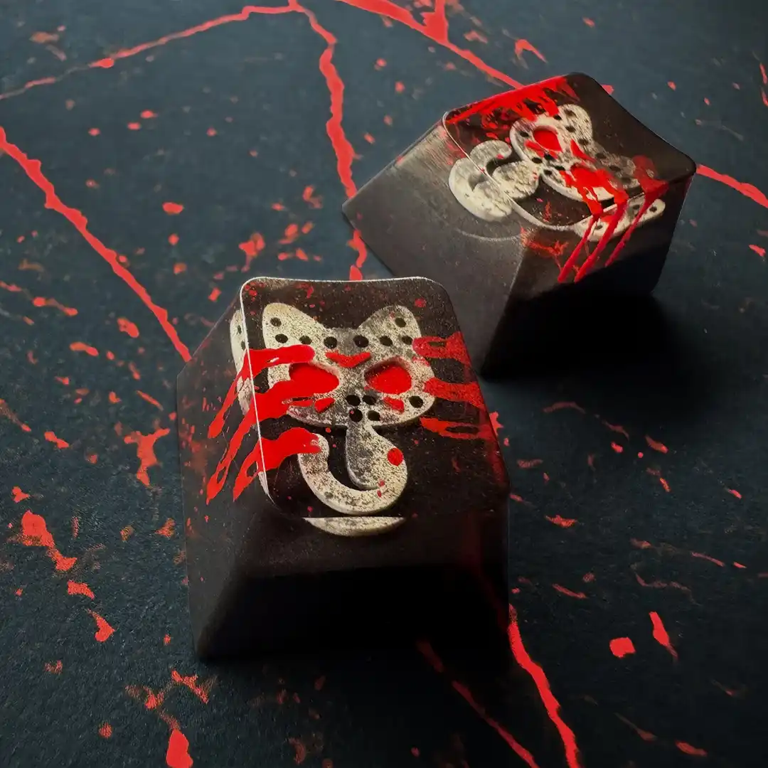 Two black keyboard keycaps with a white cat face design are covered in red splatters resembling blood, set against a dark…