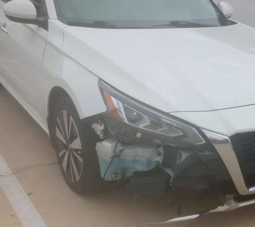 A white car has sustained visible front-end damage, with its left headlight assembly shattered and the engine bay exposed…