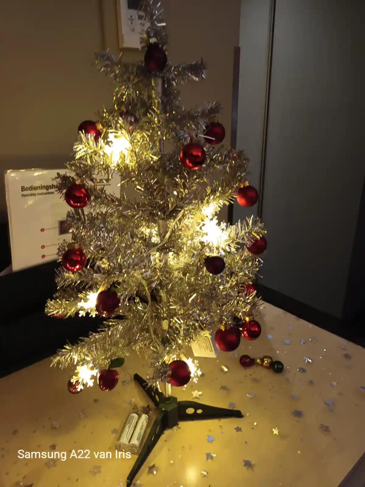 A small, silver tinsel Christmas tree decorated with red ornaments and warm white lights sits on a table covered with silver…