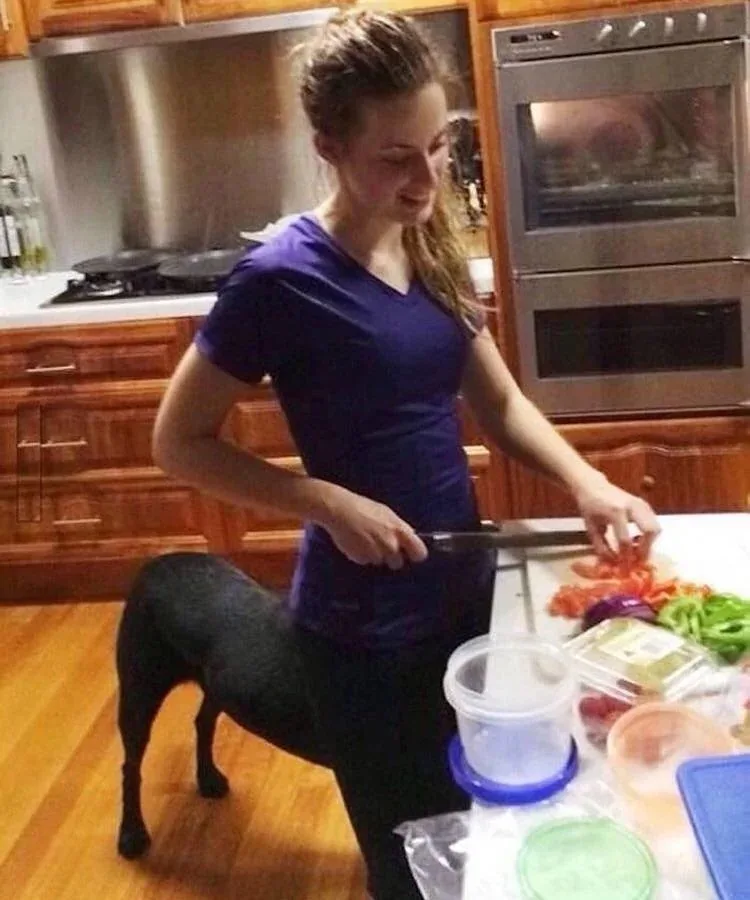 A woman in a purple shirt stands in a kitchen, holding a knife and cutting vegetables on a counter. A black dog is…