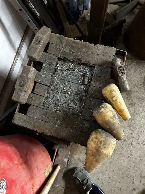 A top-down view of a worn wooden anvil block with metal hammers and chisels scattered around it in a cluttered workshop. The…