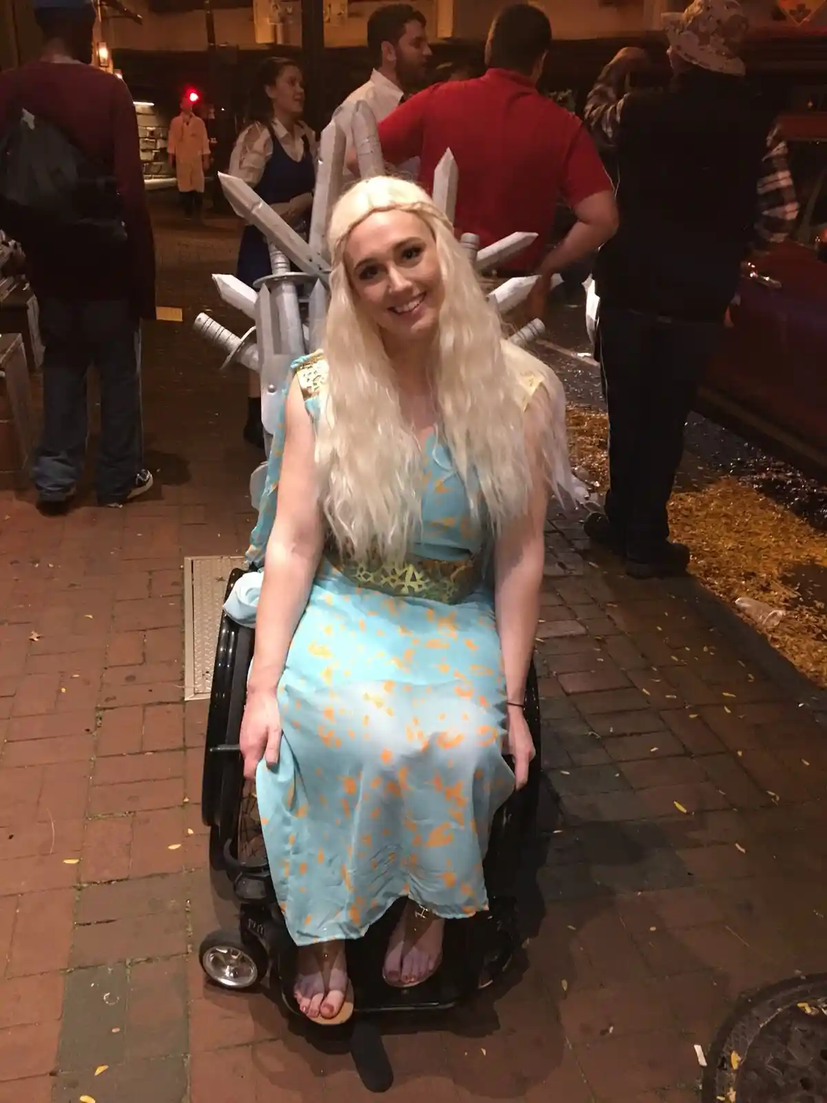 A woman with long blonde hair, wearing a light blue and gold dress, sits in a wheelchair outdoors at night. Behind her, a…