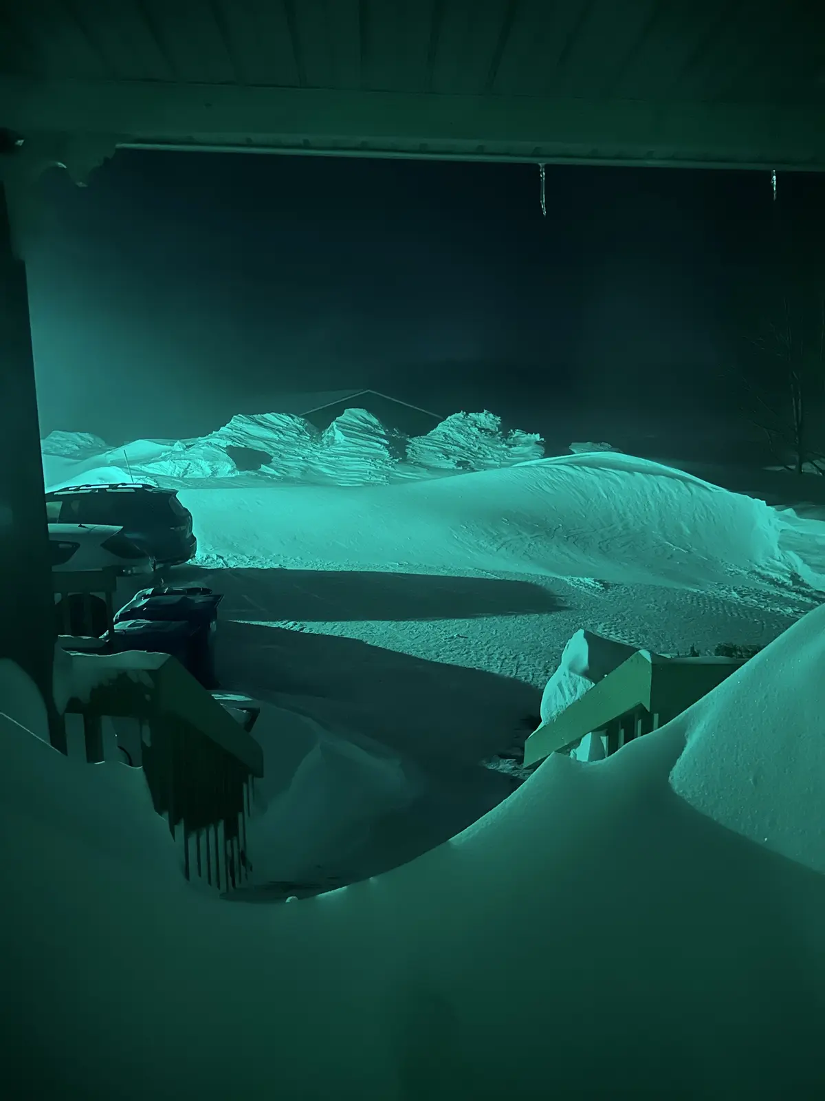 A nighttime photo taken from a porch shows a driveway and yard completely buried under deep, sculpted snowdrifts, with…