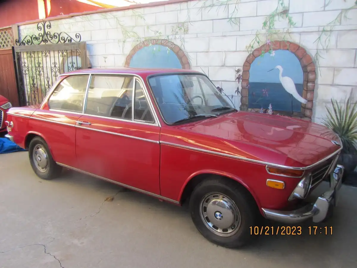 A red vintage BMW sedan is parked in front of a painted wall featuring two arched murals. The right mural depicts a white…