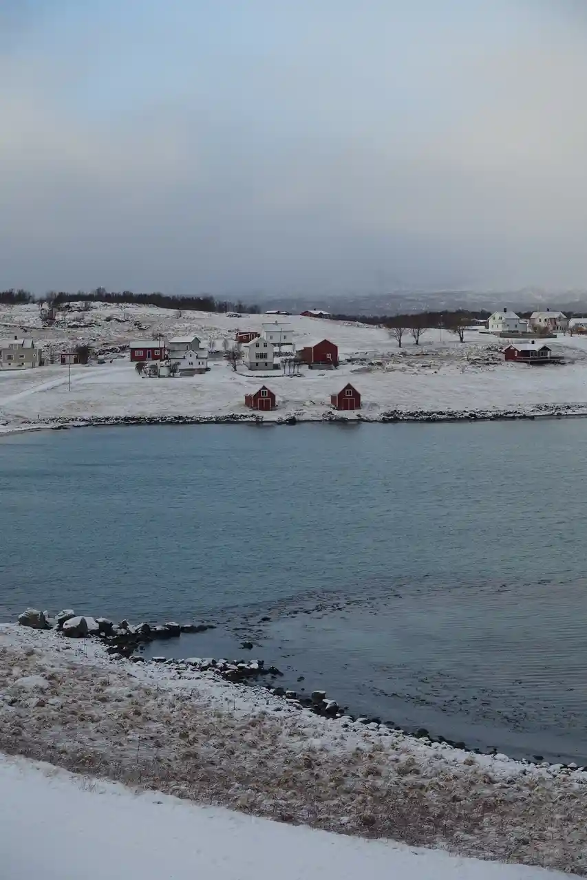 A serene winter landscape shows a snow-covered coastal village with red and white houses nestled along a calm blue waterway…