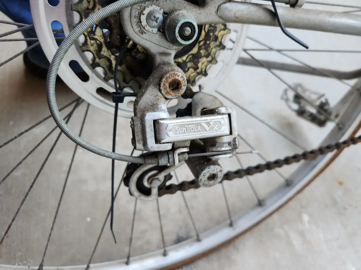 A close-up photo of a rusty, worn-out bicycle rear derailleur and gear cassette, showing the brand name 'SunTour VX'…