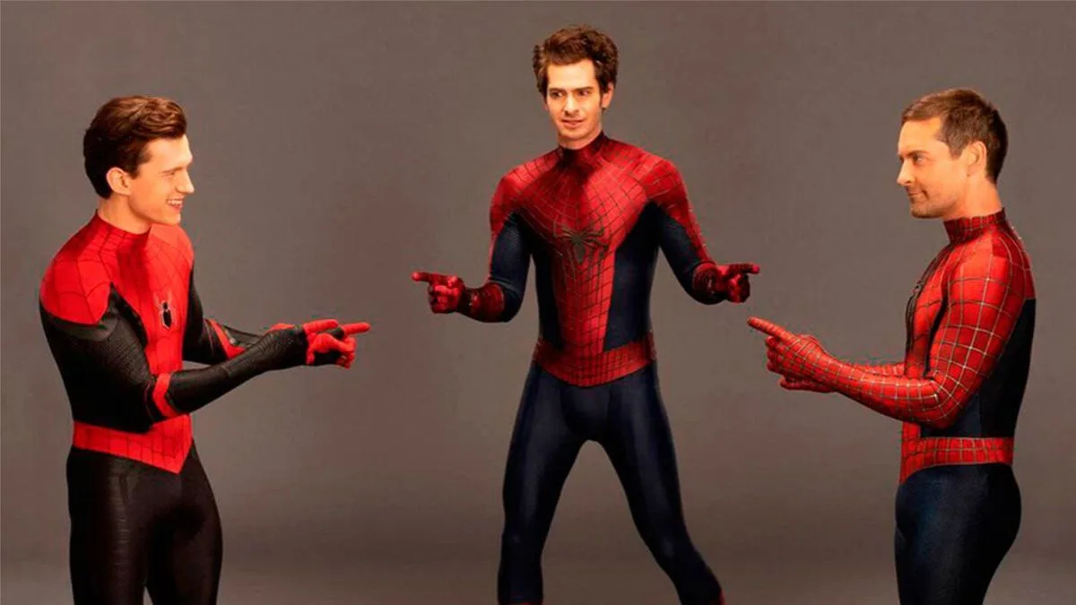 three men in different Spider-Man costumes point at each other against a gray background. The man in the center wears the…