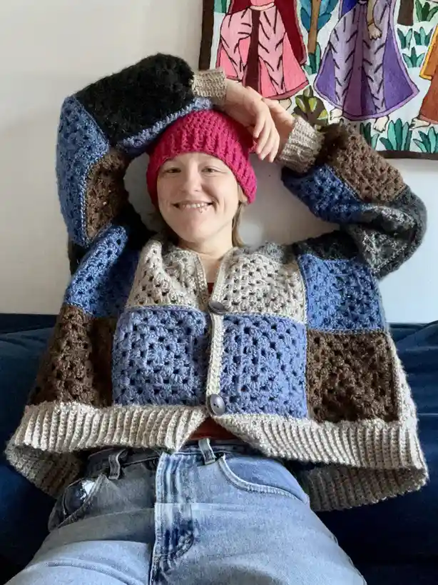 A smiling person wearing a colorful, patchwork-style crocheted cardigan with a pink beanie, posing on a couch with hands…
