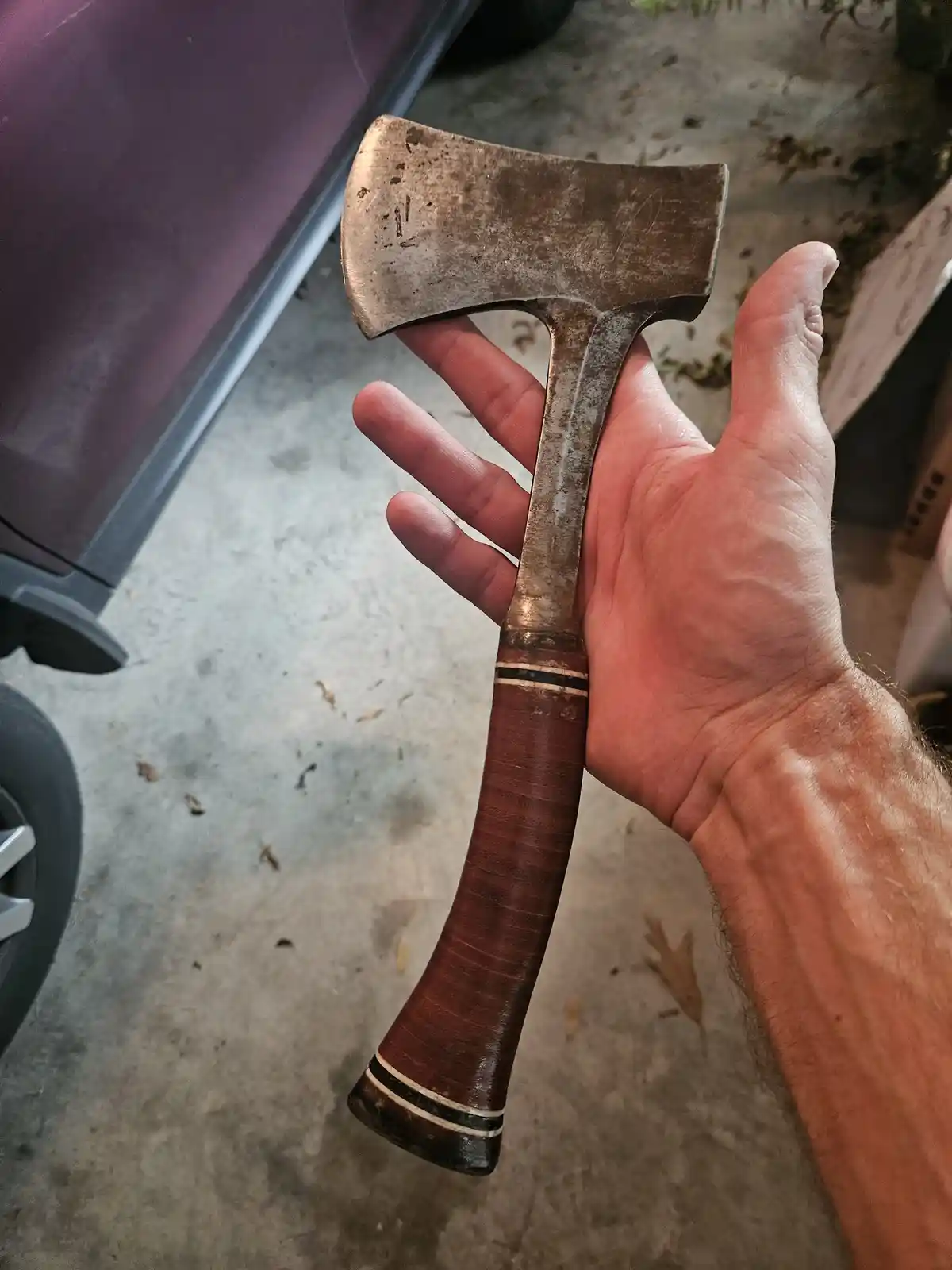 A person's hand holds up a well-worn axe with a brown leather-wrapped handle and a metal head showing signs of use. The…