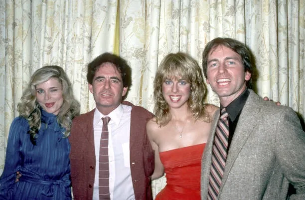 A vintage photo of four people posing together in front of patterned curtains. From left to right: a woman in a blue dress…