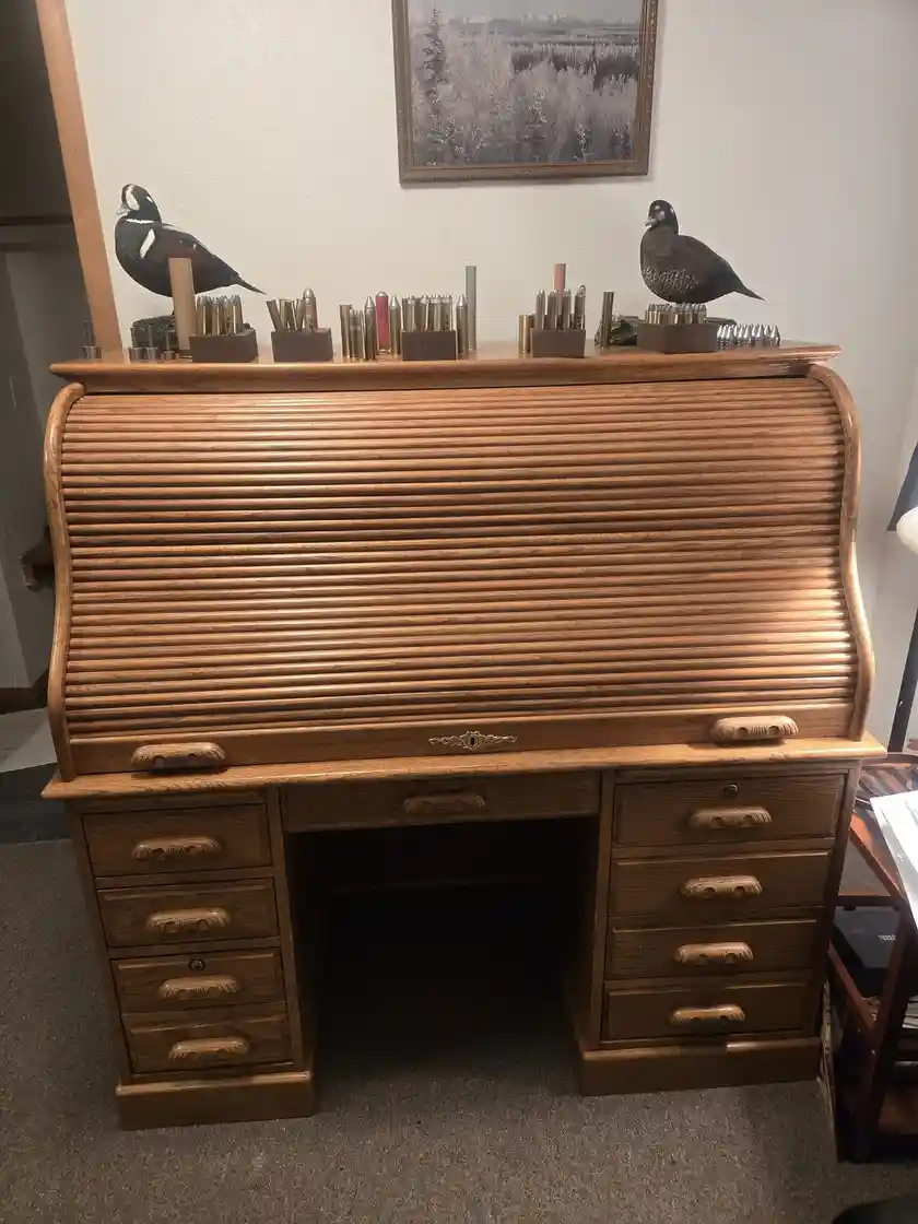 A wooden roll-top desk with a closed shutter, topped with two taxidermy ducks and multiple displays of rifle cartridges…