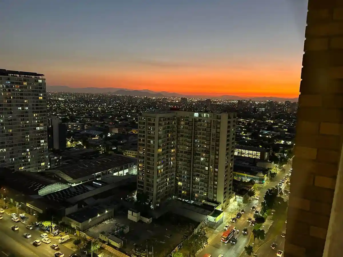 A high-angle cityscape photo at dusk, showing a vibrant orange sunset over distant mountains, with numerous lit-up apartment…