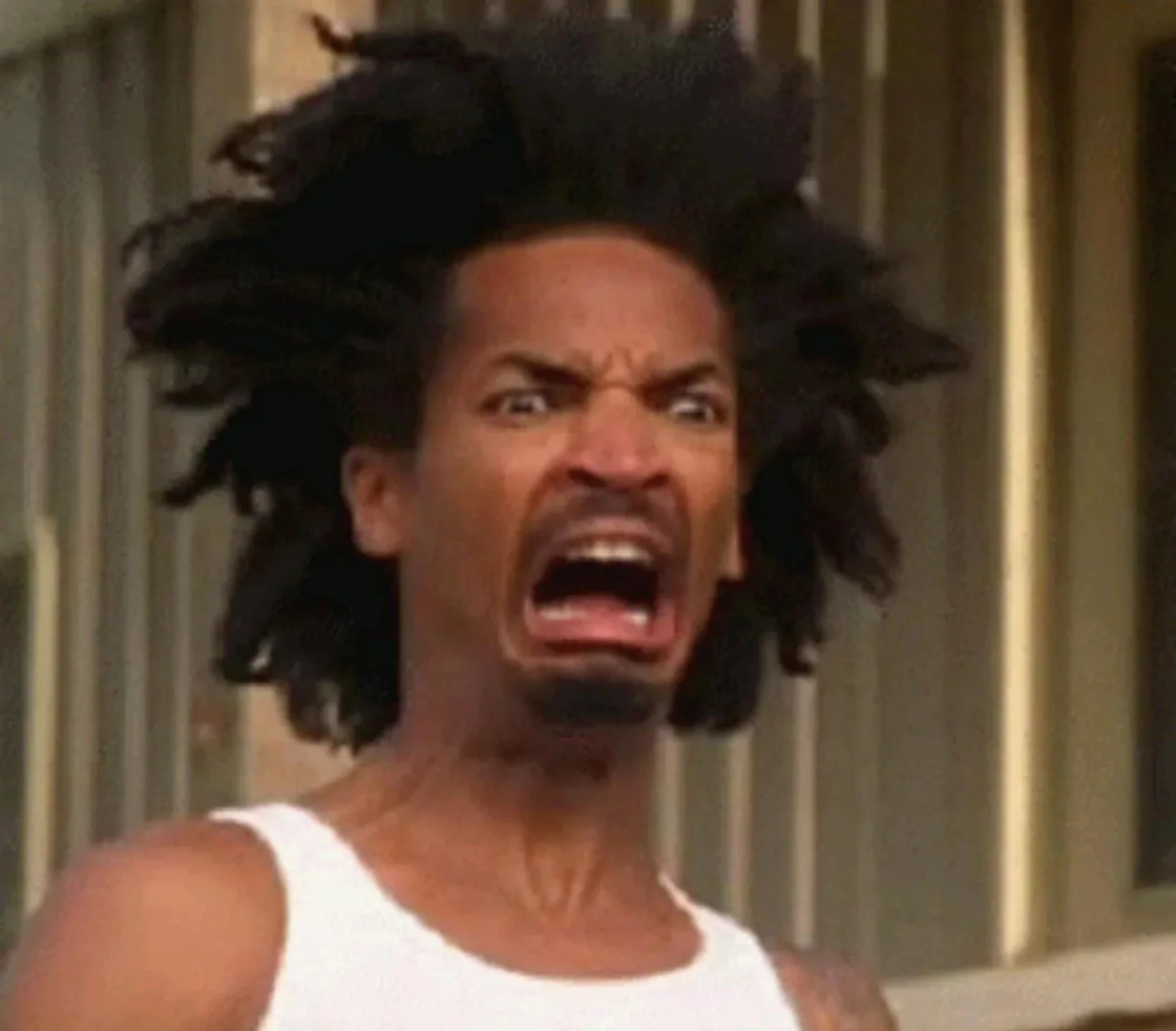 A man with wild, dark dreadlocks and a white tank top is captured mid-shout with his mouth wide open, eyes bulging, and…