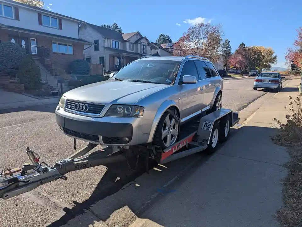 A silver Audi station wagon is loaded onto a U-Haul flatbed trailer parked on a residential street. The car is positioned…